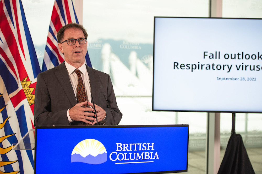 Adrian Dix, a middle-aged white man with short brown hair, speaks from behind a podium. The podium is blue and says ‘British Columbia.’ Behind Dix, there are two BC flags.