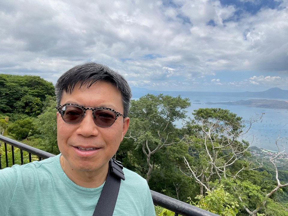 A person wearing black sunglasses and a seagreen T-shirt takes a selfie in front of a blue sky and hills.