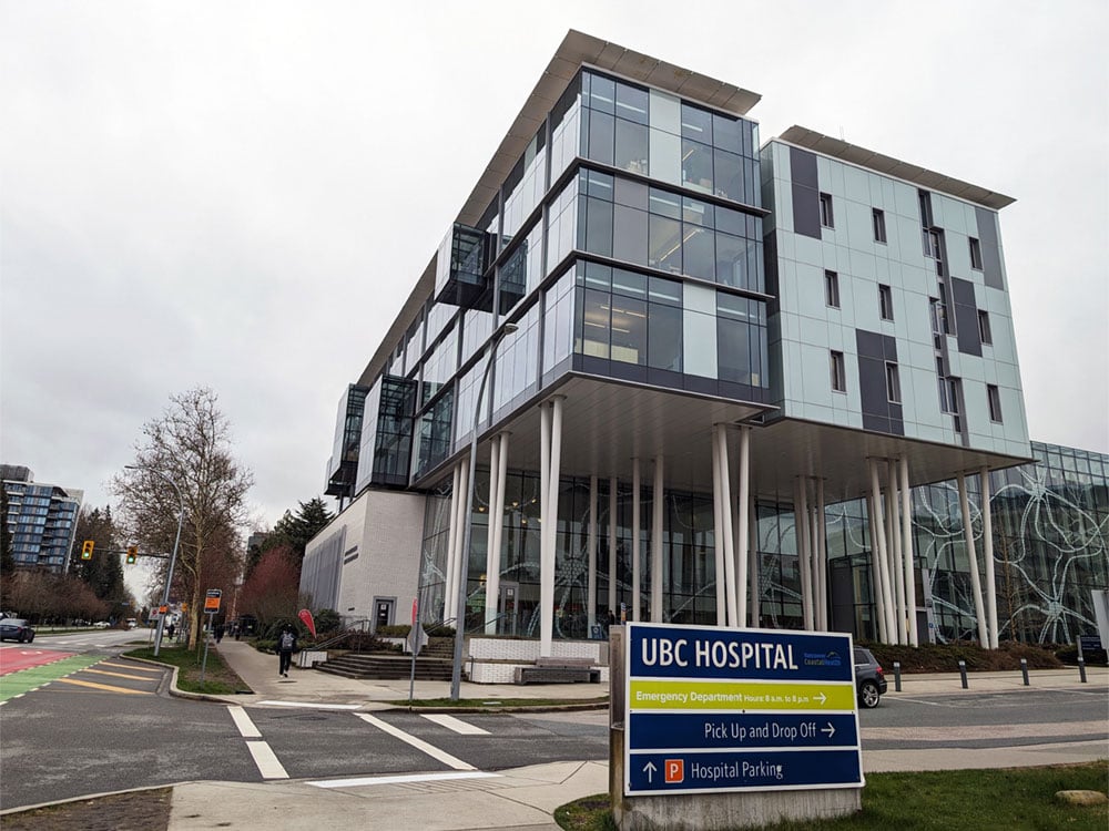 The UBC Hospital on an overcast day. A blue sign in front of a modern building with tall pillars reads, ‘UBC Hospital. Vancouver Coastal Health.’
