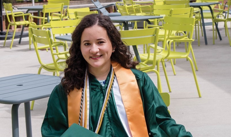 A young woman with dark curly hair and light skin wearing a graduation robe smiles while seated at an outdoor table.