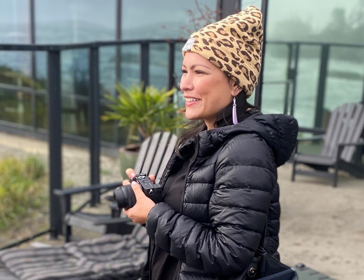 A light-skinned woman with long black hair, wearing a leopard spotted cap and holding a camera, smiles and looks to the left of the frame.