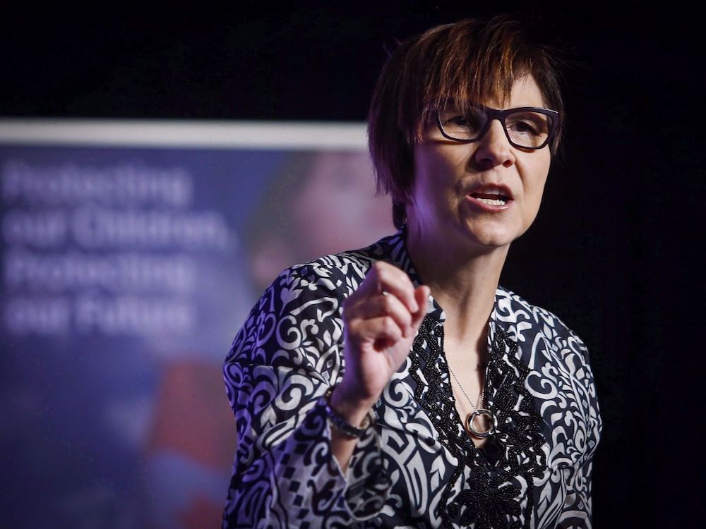 A middle-aged woman with medium-light skin tone, dark short hair and black-rimmed glasses gestures while speaking.