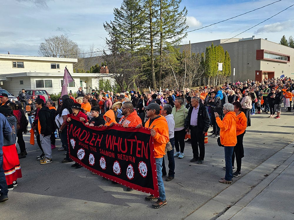 A crowd of people stand on a residential street, many of them wearing orange to represent support for residential school survivors.