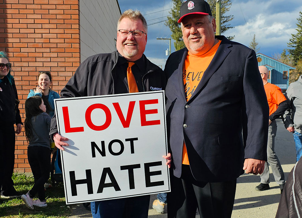 Two men stand side by side holding a sign that says 'Love not hate.' The men are wearing black and the man on the left is wearing an orange tie while the man on the right wears a ball cap and an orange T-shirt.