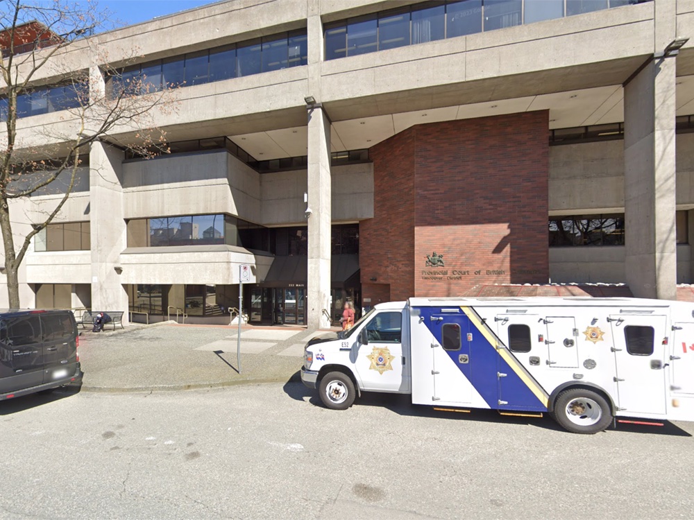 A looming, three-storey concrete building. A prison transport vehicle is parked out front.