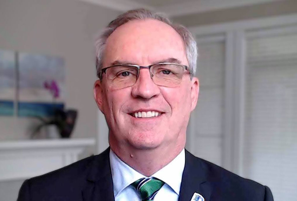 A light-skinned man with short silver hair and glasses smiles at the camera. He wears a blue jacket, white shirt and green striped tie.