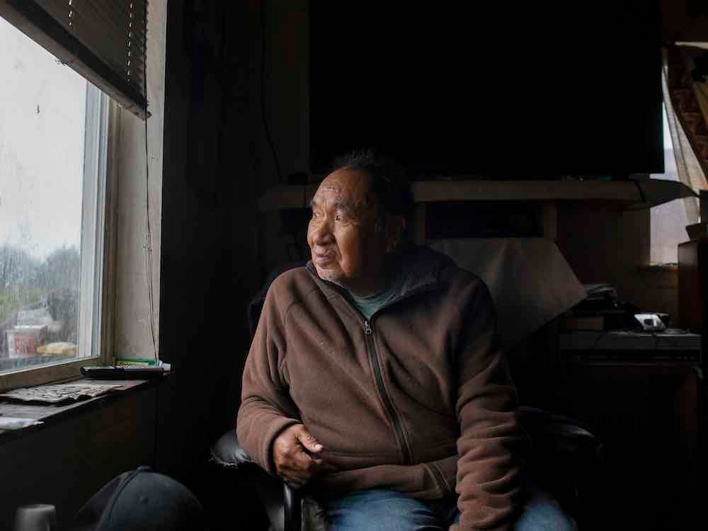 An elderly Indigenous man with medium skin tone looks to the left of the frame, out the window of his home. He is sitting in a dimly lit room wearing a brown zip-up hoodie.