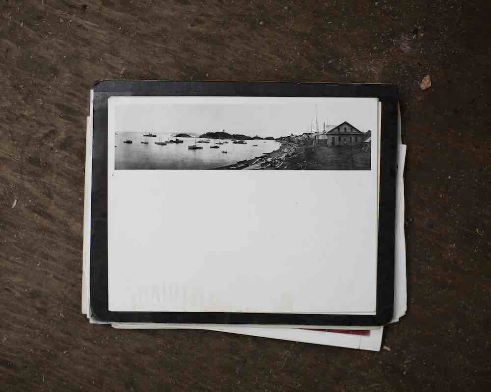 A photograph, shot from above, of a black and white photo depicting a panoramic view of Yuquot Bay and the fishing boats in the water. The photo sits atop a small stack of other paperwork on a dark brown tabletop.