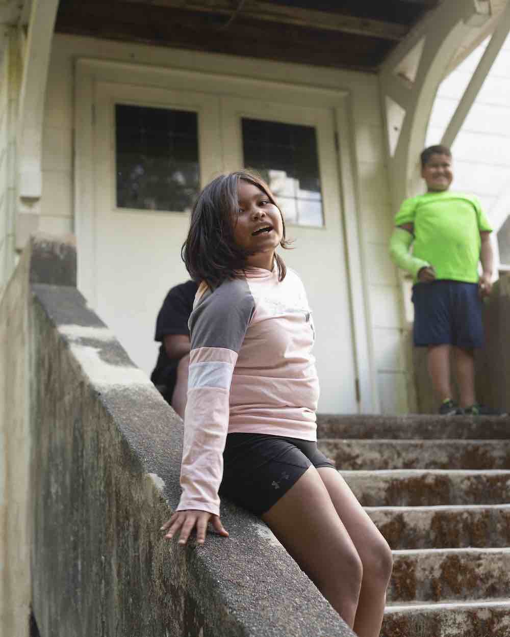 Three children stand on the front concrete steps of a white building. The girl in the foreground has short dark hair, medium skin tone and a light pink shirt with black athletic shorts. A boy in the background is standing near the door behind her wearing a neon-green T-shirt.