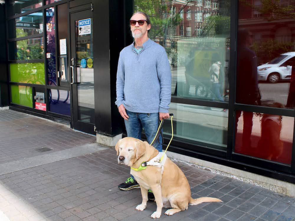 A light-skinned man with short hair and a tidy grey beard stands on a sidewalk. He wears jeans and a blue sweater and holds the harness of a large golden-coloured dog.