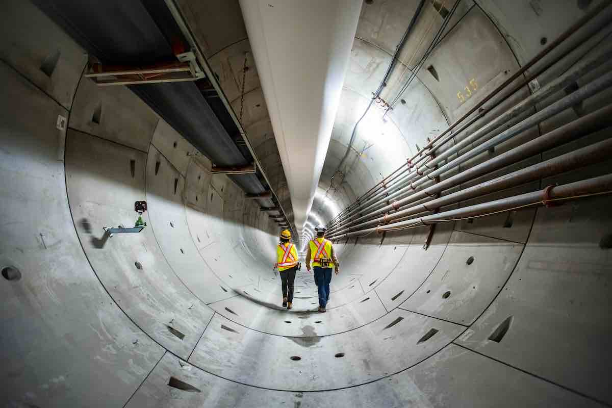 Two workers in fluorescent yellow construction hats and white helmets walk with their backs to the camera in a large grey concrete tunnel. A large white pipe spans across the top of the tunnel and several thinner pipes run to the workers’ right.