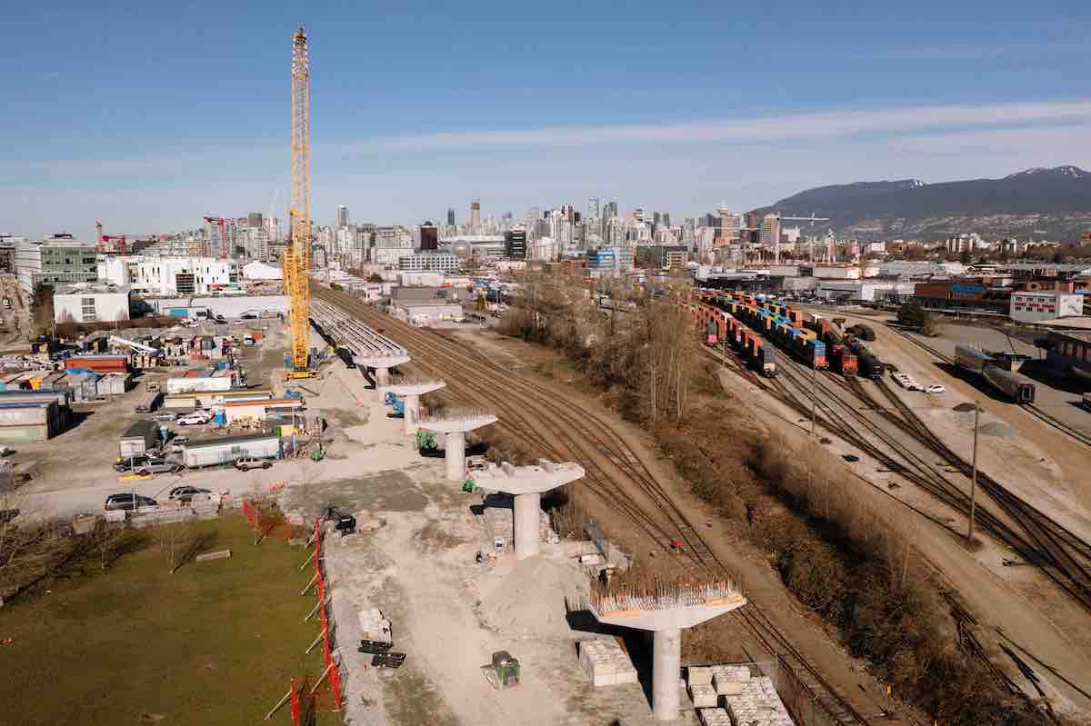 An elevated view of a row of several raised concrete platforms for the future Broadway Subway line that run alongside train tracks to their right. A yellow construction crane is to the left of the platforms. It’s a bright winter day.