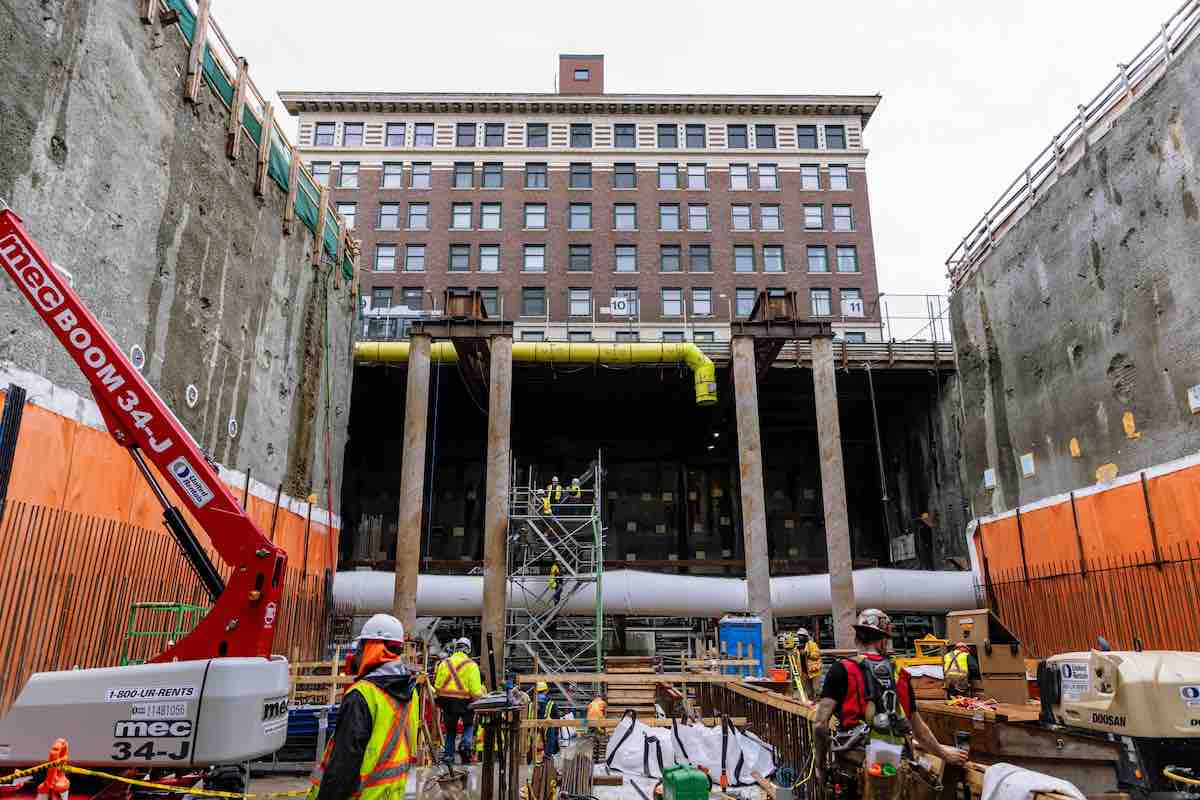 An underground construction site is flanked by two steep concrete walls on both sides, with yellow horizontal piping running across the back wall flanked by two pairs of vertical cylinders. Higher up in the background, an older brown brick building looms against a white sky.