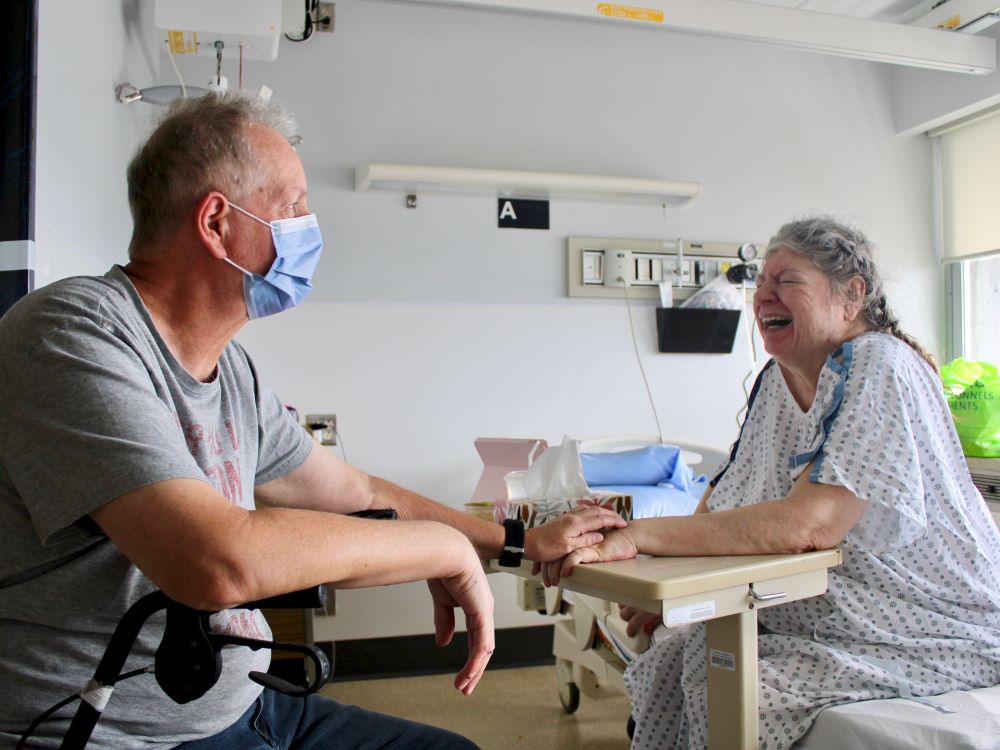 Two older people sit in a hospital room. A man on the left wearing a surgical mask reaches out and rests his hand on the hand of a grey-haired woman in a hospital gown who is laughing.