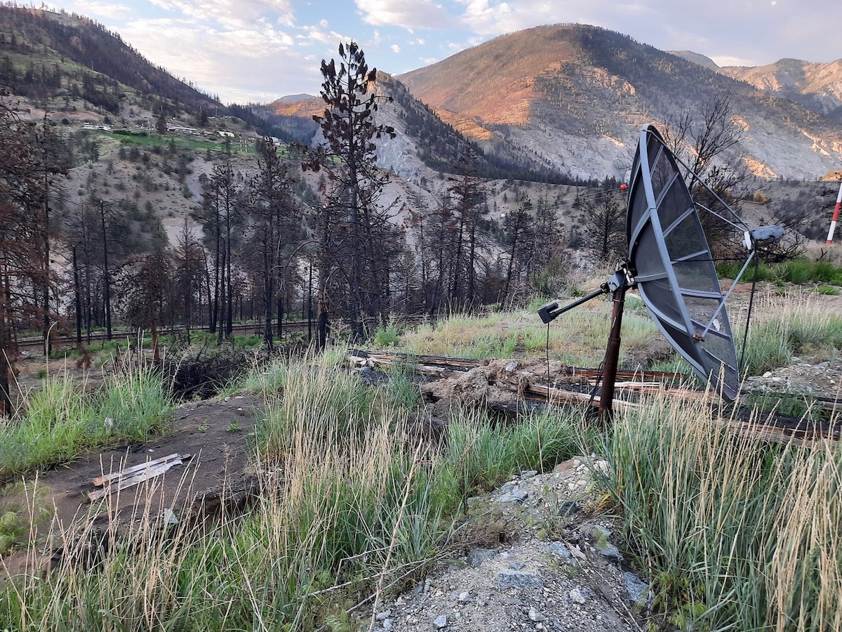 In the foreground is a grassy area with gravel and other debris, some appearing burned, on the ground. There is also a satellite dish. There are blackened trees and mountains behind.