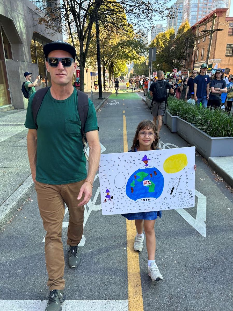 A young girl with long brown hair and glasses walks in a city bike line holding in front of her a poster board illustrated with a child's drawing of the Earth. Beside her walks her father, wearing a baseball cap and sunglasses. In the background is a crowd of people holding up signs.