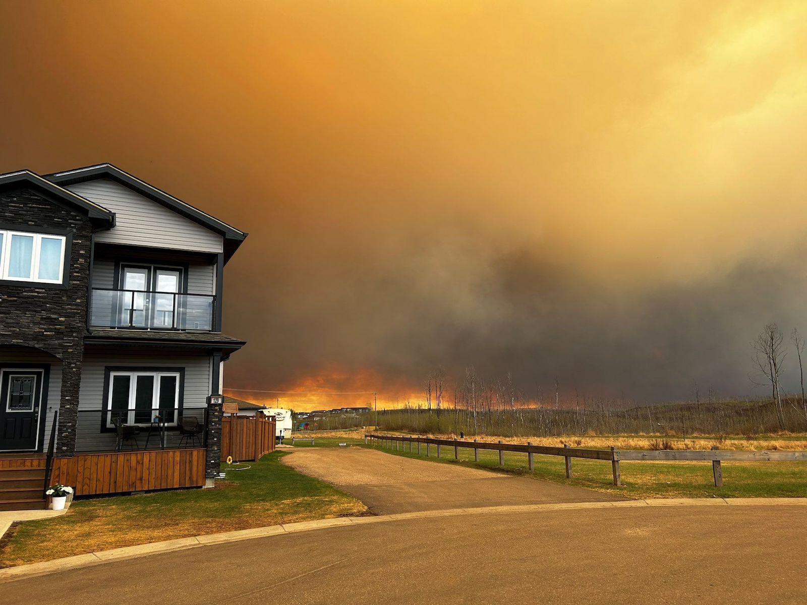 A home sits next to a field showing the scorched remnants of trees, under a smoke-filled sky tinged orange from the glow of fires visible in the background.
