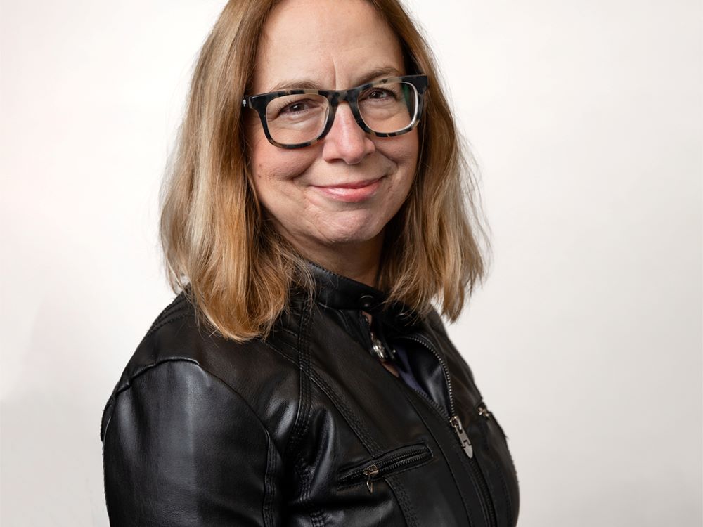 A person with light skin and shoulder-length hair stares directly at the camera in front of a white background. They are wearing a black leather jacket and glasses with matte, square frames.
