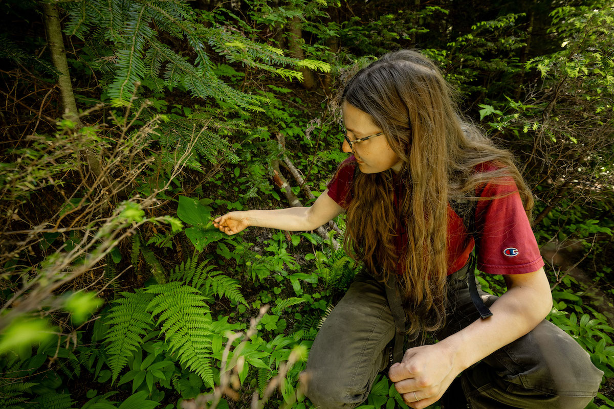 A younger woman with light skin tone and dark brown hair crouches down in the forest. She is wearing a red shirt and green khaki pants.