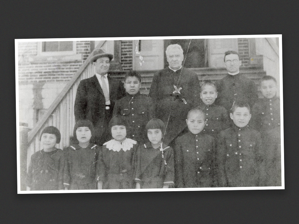 A black and white photo shows 10 Indigenous children in uniforms standing with three men, two in priests’ clothing, outside a brick building.