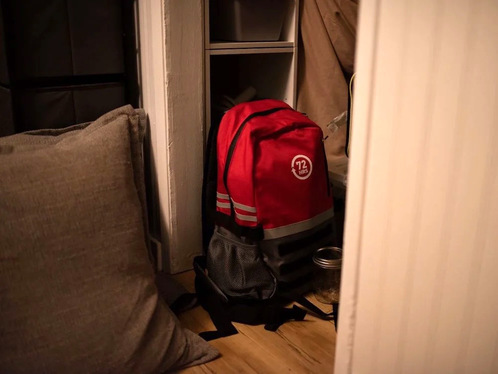A red and black backpack with a '72 hrs' logo sits on the floor of a home.