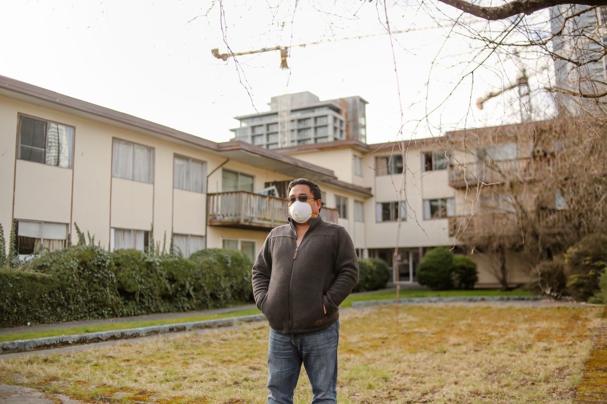 A middle-aged man in a white medical mask stands with his hands in the pockets of his grey jacket, which he wears with blue jeans. He is standing in front of a beige three-storey apartment complex.