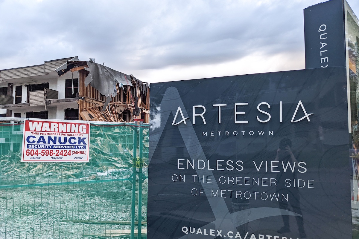 A green construction fence rises in front of a demolished white three-storey apartment building. In the foreground, a navy real estate sign advertises a new development called Artesia.