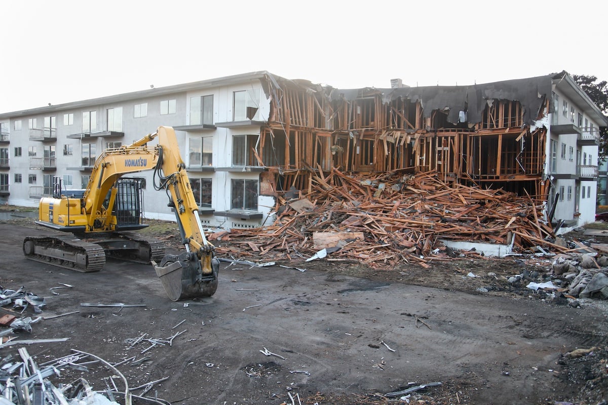On the left, a yellow pile digger stands before a white low-rise apartment building in the process of being demolished. Broken wood is visible across the building on the right and its facade is visible on the left.