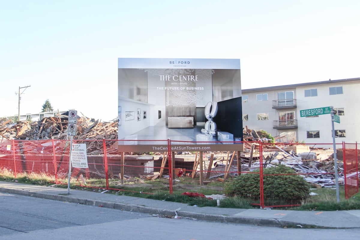 A black and white real estate ad reading 'The Centre' stands behind a construction fence amongst rubble from a demolition. A beige low-rise apartment building stands to the right of the frame.