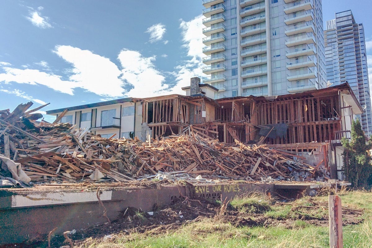 A beige low-rise wood-frame apartment building stands partially demolished behind a large pile of broken wood.