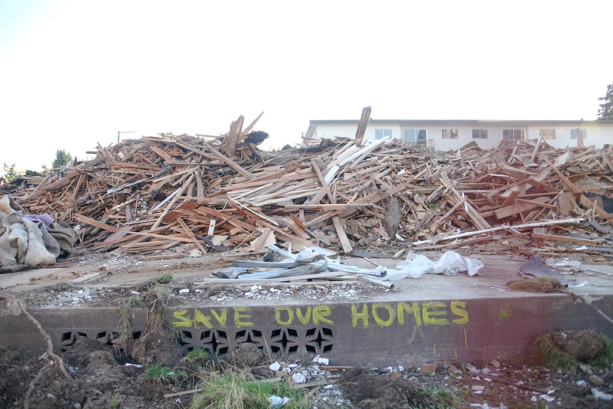 A pile of wood from a demolition sits atop a concrete fence. In yellow handwritten text, the phrase 'Save our homes' can be seen across the top of the concrete.