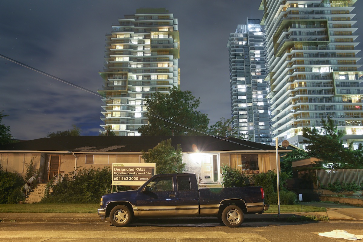A blue pickup truck is parked outside a one-storey row of bungalows behind a sign that reads 'Designated RM5s High-Rise Development Site.' Behind them loom contemporary highrise condo towers. It’s nighttime.