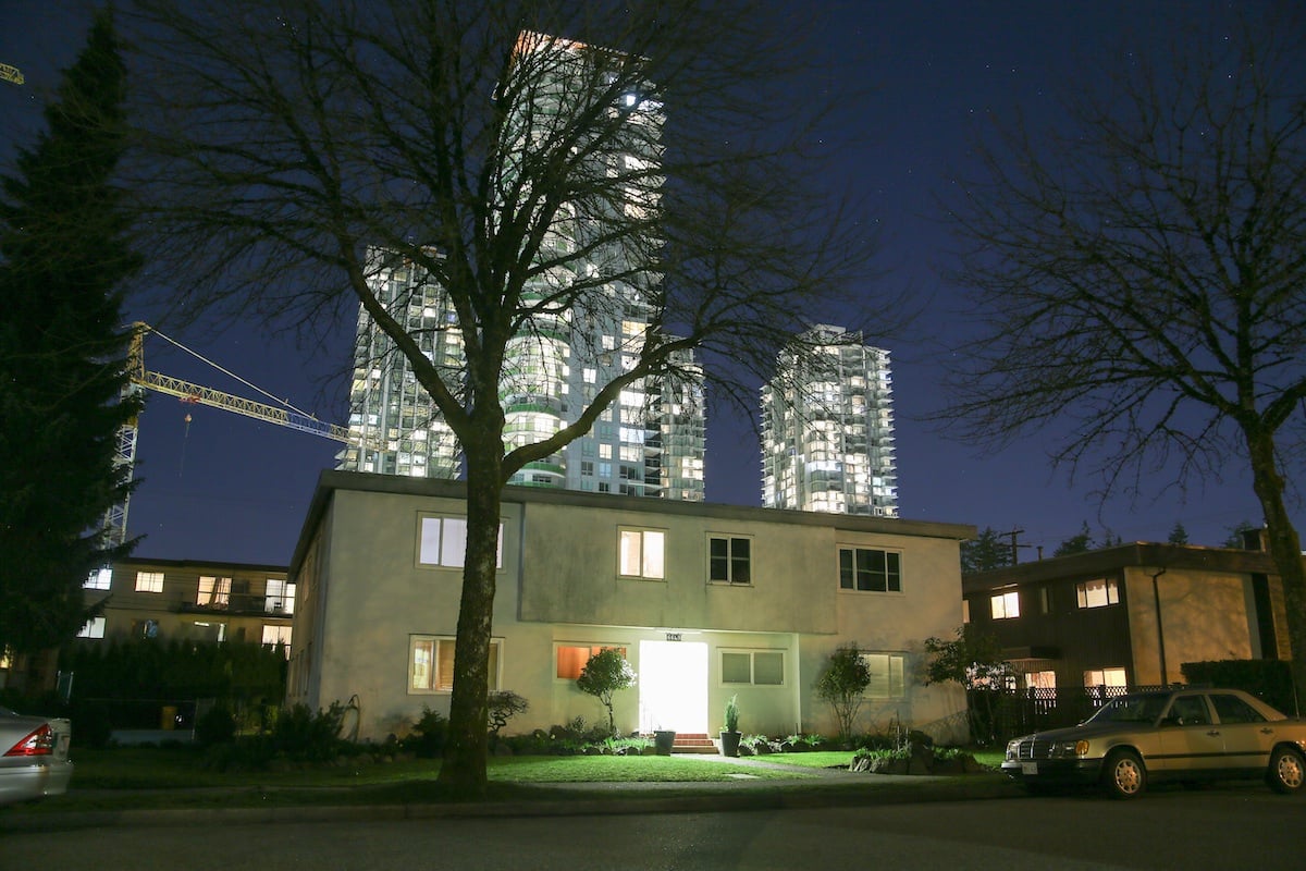A white two-storey apartment building’s door glows white in the night. Behind it are construction cranes and contemporary highrise condo towers.