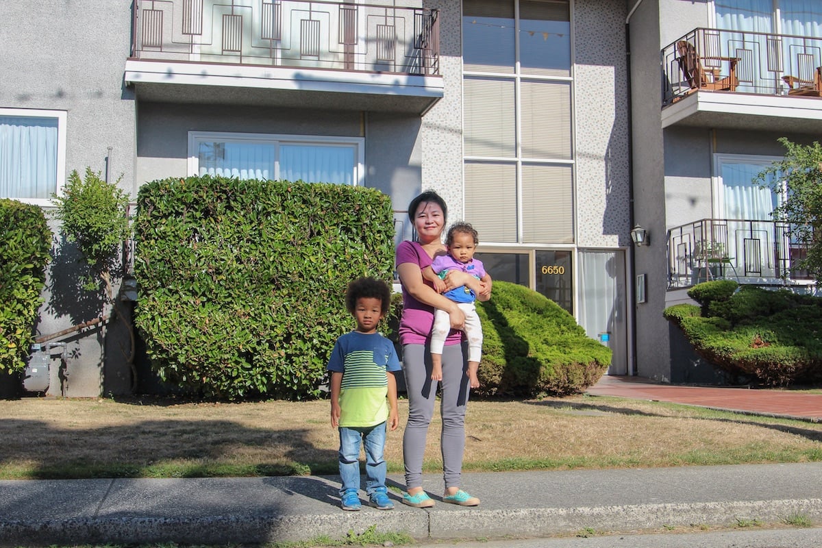 A woman with dark hair tied up, light skin, a dark pink T-shirt and grey leggings stands on a sidewalk holding a toddler and beside a young school-aged child with medium skin and curly hair. They are in front of a grey low-rise apartment building.