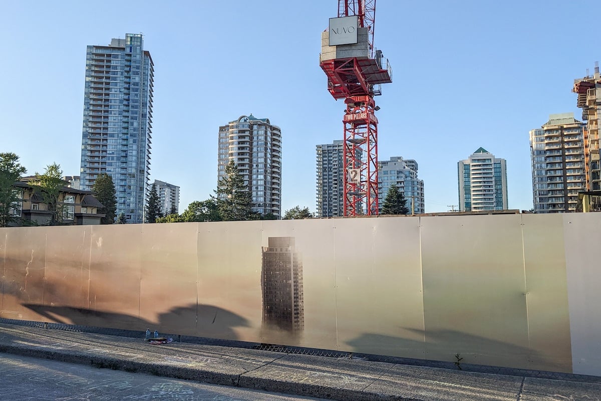 A photo of a construction fence featuring a large wraparound ad for real estate features a highrise tower against a hazy orange sky. Behind it are construction cranes and towers.