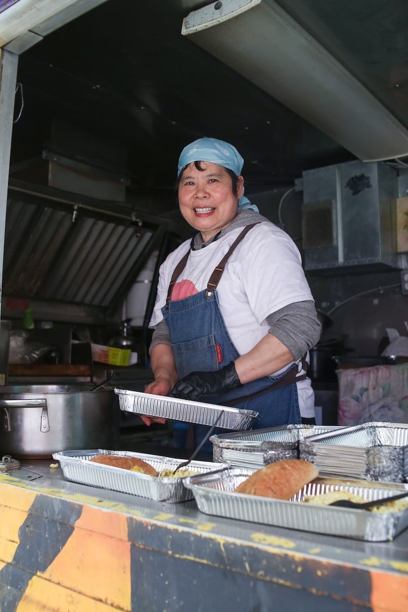 Anna Lao has short dark hair tied back with a light blue headscarf. She wears a navy blue apron over a white T-shirt. She is holding a large foil pan and standing in a commercial kitchen.