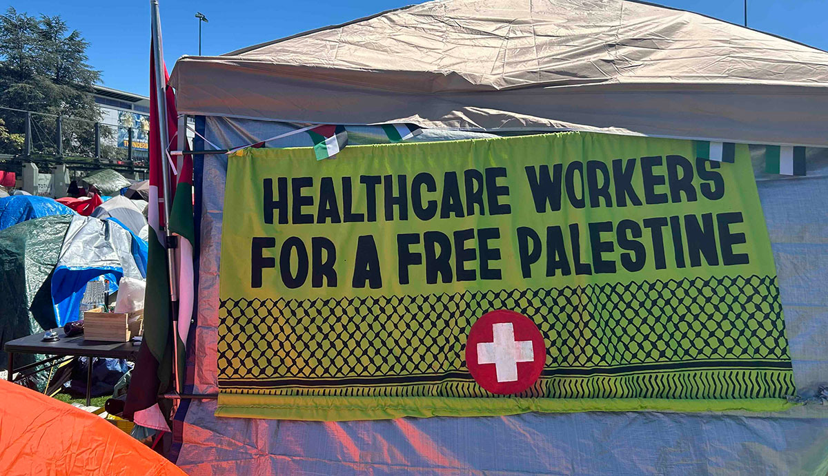 A large green handmade sign on the side of a beige tent reads 'Healthcare workers for a free Palestine.'