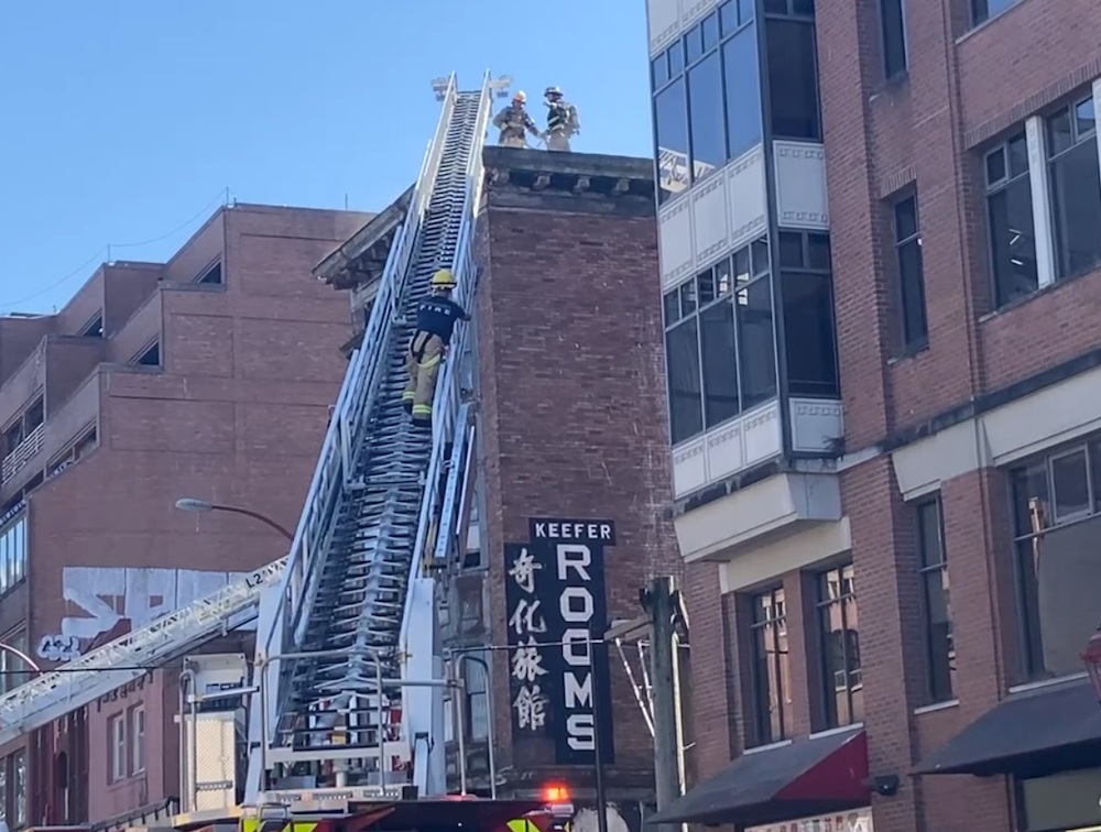 A white firefighter ladder extends up to the roof of a brown brick building. The side of its wall reads 'Keefer Rooms' in large white text with a vertical row of Chinese characters down the side.