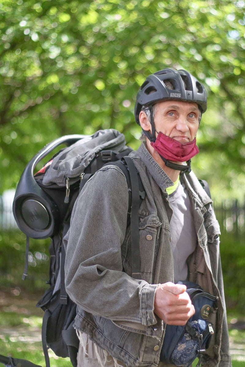 Steve Nelson stands against a lush green backdrop of bright green trees. He wears a black bicycle helmet, a burgundy fabric face mask tucked near his chin, a backpack and a dark grey jean jacket over a grey T-shirt. He has light skin and light eyes.