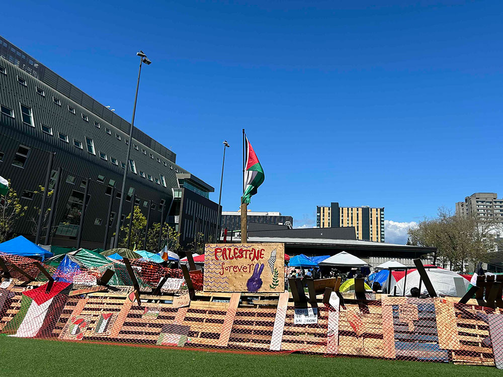 A makeshift barricade of wooden pallets covered with orange industrial mesh features a wooden placard that reads 'Palestine Forever' with a purple hand making a peace sign. Behind the barricade are tents to the right of a grey building. The sky is blue.