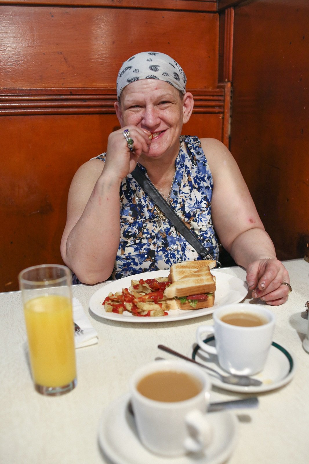 A woman with light skin, a light blue patterned bandana and a sleeveless blue and white floral top sits against a warm wood fixture at a diner table spread with small white coffee cups, an orange juice and a plate with a sandwich and salad.