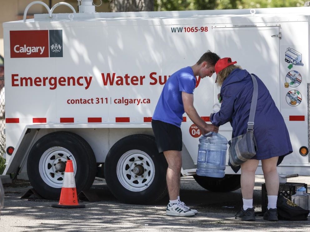 A young man and a woman handle a large clear plastic bottle of water next to a truck marked 'Emergency Water Supply.'