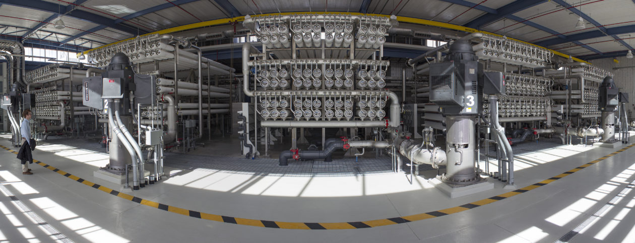 In a wide-angle view of the Kay Bailey Hutchison Desalination Plant in El Paso, Texas, an array of pipes and tubes lies behind a yellow and black striped line. A person in a blue shirt stands off to the left of the image.