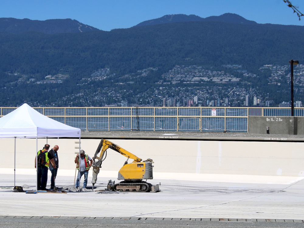 Three people in visibility vests stand next to construction machinery boring into concrete inside an empty outdoor pool. The North Shore can be seen in the bakcground across Burrard Inlet.
