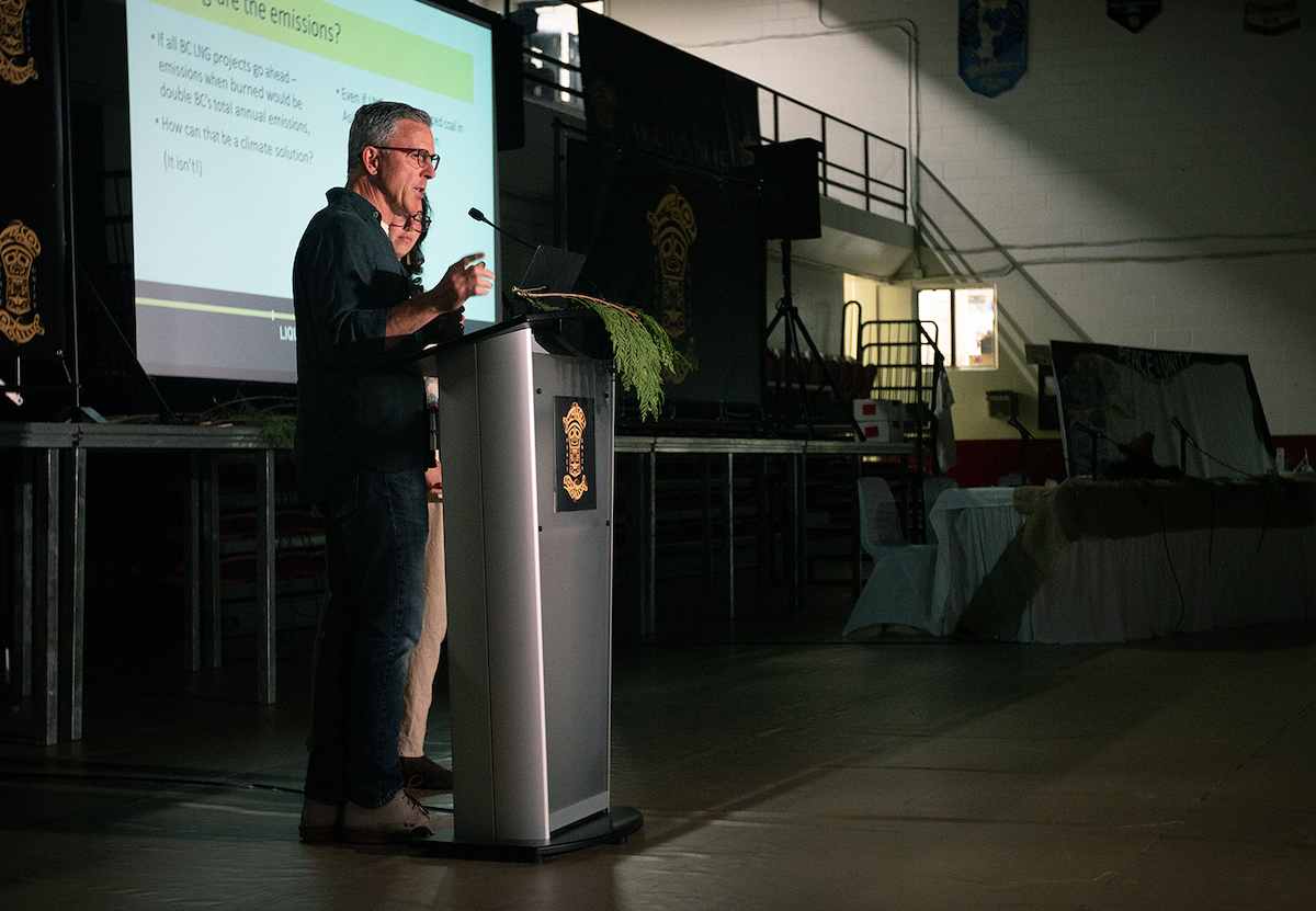 A man with glasses, a fair complexion and greying hair speaks at a podium. There is a screen with a PowerPoint presentation behind him.