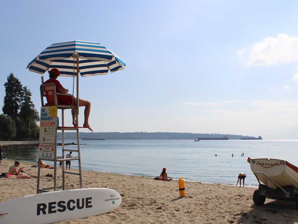 A lifeguard wearing a red T-shirt and shorts sits in a lifeguard chair, with a rescue board at the base of the chair, and a wooden rescue rowboat a little ways away in the sand. Kids and adults sit on the beach, and play in and around the water.