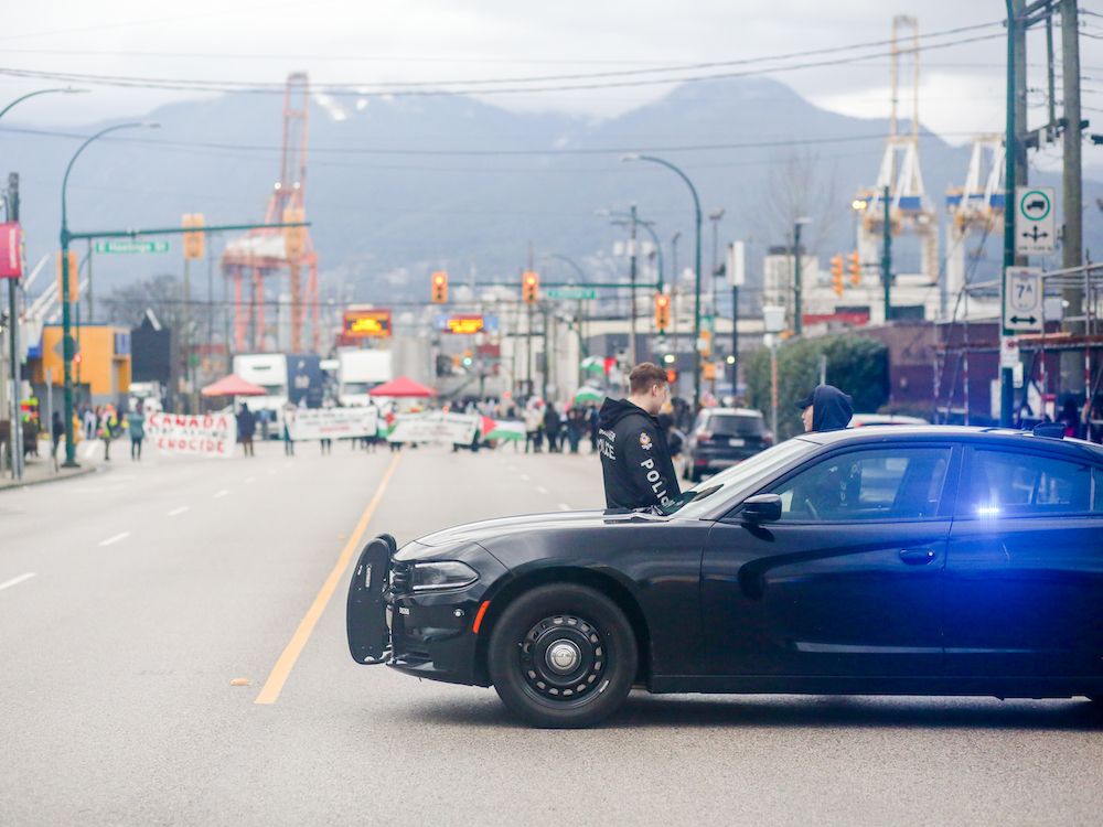 In the foreground, two police officers stand beside a police car. In the background, pro-Palestinian protesters carry placards and Palestinian flags in front of the port in Vancouver.