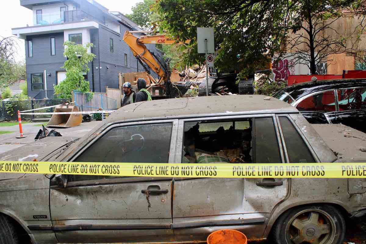 A burned, battered car sits behind yellow police tape. In the background are a yellow backhoe and building rubble.