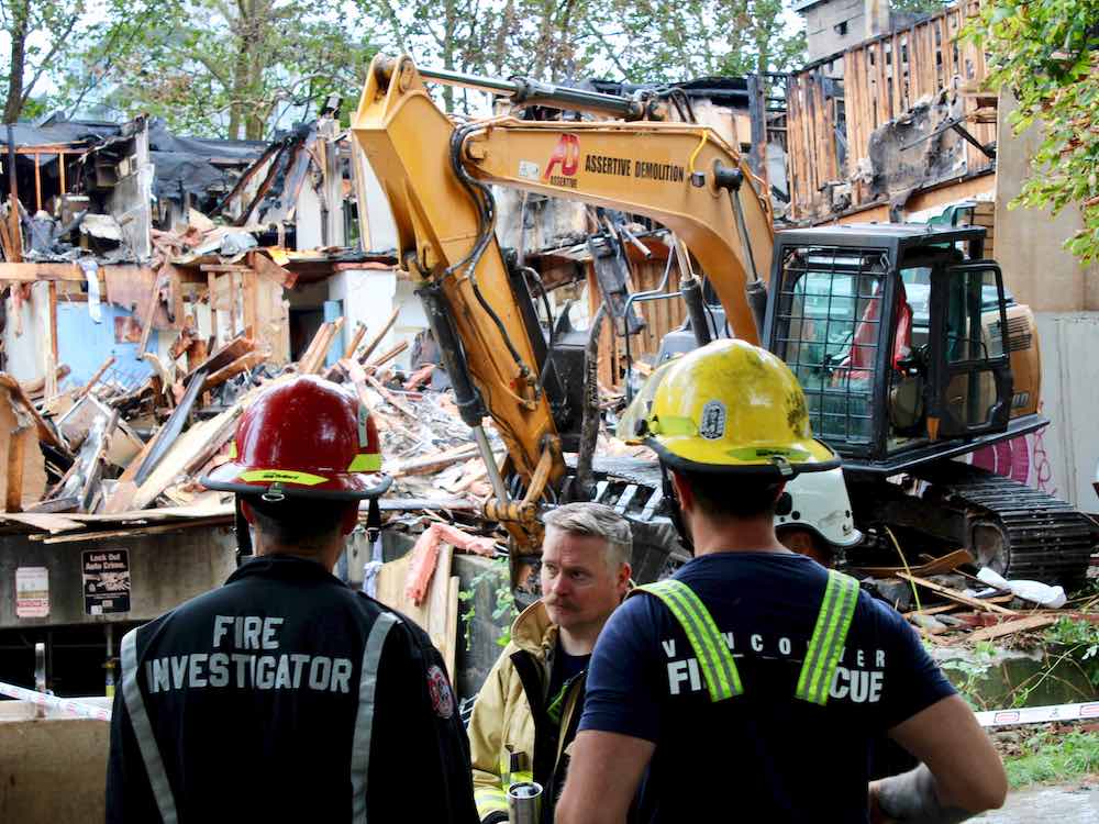 A large backhoe stands in the rubble of a burned apartment building. In the foreground three firefighters stand watching the demolition.
