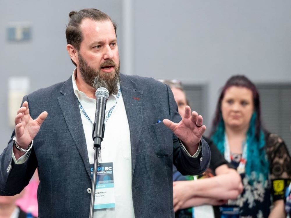 A light skinned man with brown hair pulled in a ponytail, a brown and grey beard, and wearing a charcoal sport coat speaks into a standing microphone.
